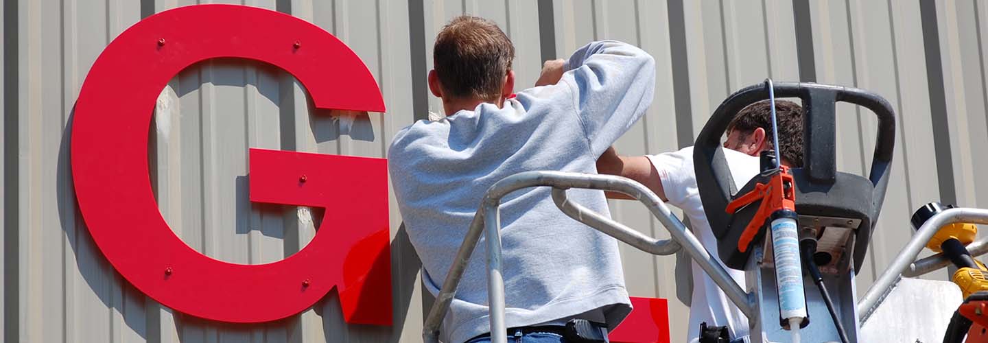 A person installing signage on a building, using a ladder and tools, with a clear blue sky in the background.