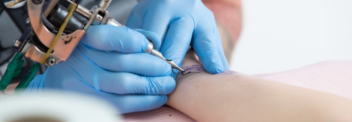 A close-up of a tattoo artist's hand holding a tattoo machine over a client's arm, tattooing a design.