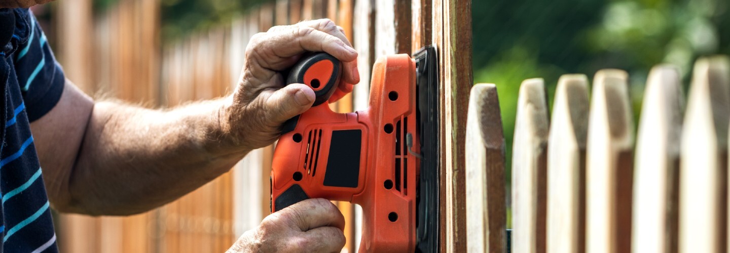 A picket fence being sanded, with green grass and blue sky in the background.