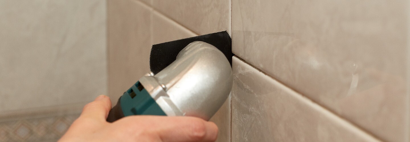 A person is regrouting tiles in a bathroom, using a tool to remove old grout.
