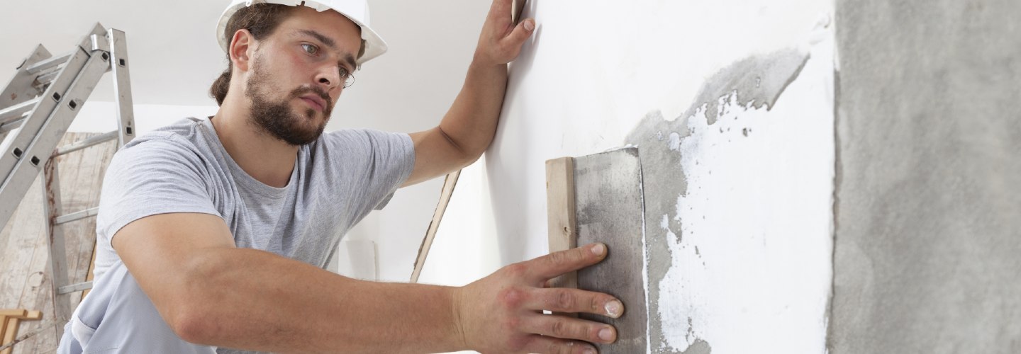 A close-up photo of a person repairing a plaster wall, using a putty knife to apply plaster compound to a damaged area.