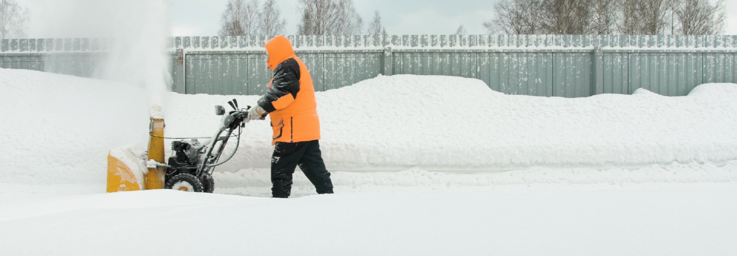 A worker using a snowplow machine clearing a snowy road during winter, with snowflakes falling and a white landscape in the background.