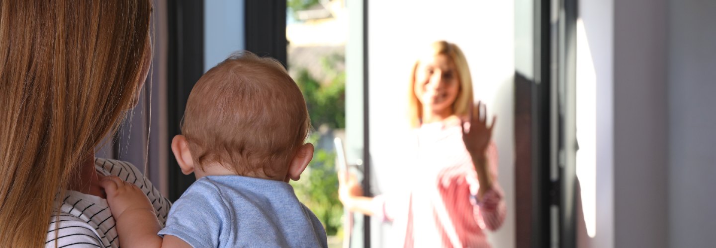 A baby being carried by a nanny, saying goodby to their mother as she walks out the door.