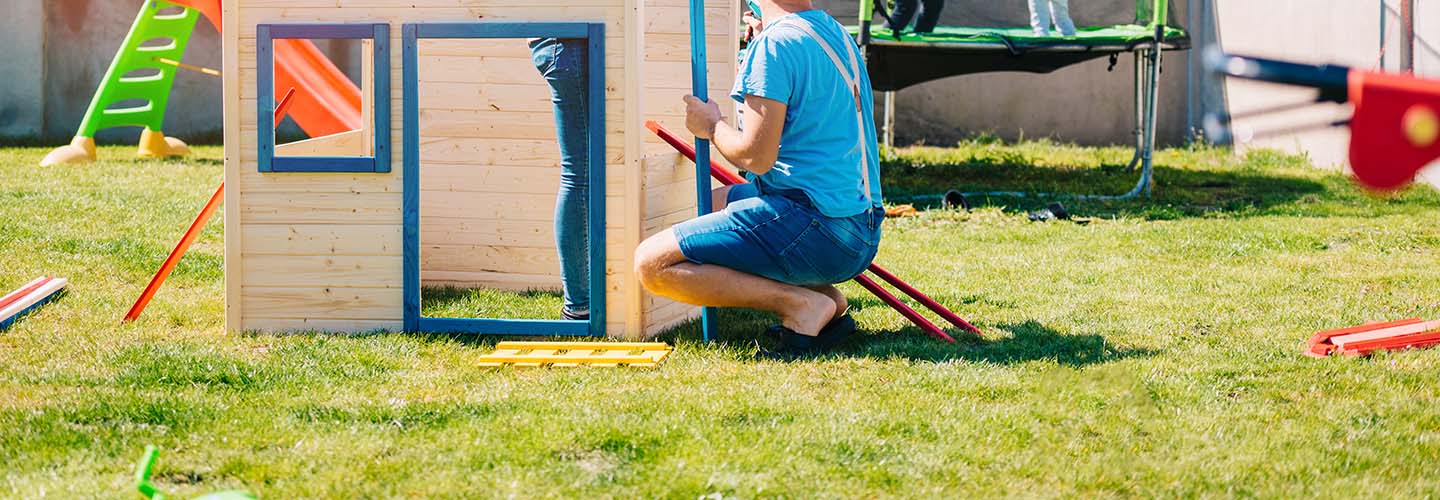 A couple of people building a playground in a sunny backyard with lush green grass.