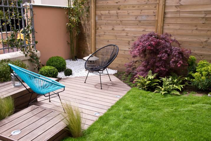 A cozy backyard setup featuring a wooden deck with colorful blue and black chairs, vibrant plants, and a neatly landscaped garden with a wooden fence