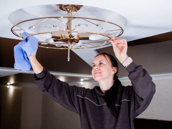 woman happily wiping dusty metal light fixture, chandelier