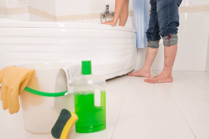 Bathtub cleaning materials on bathroom floor, barefoot man cleaning bathtub