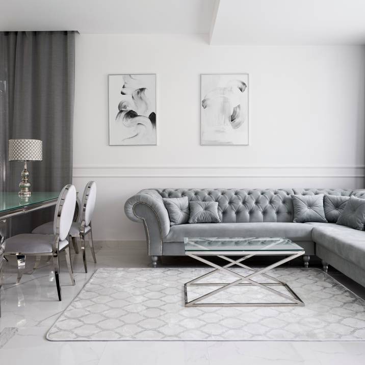Luxurious and glamorous living room interior with silver and gray color scheme. Quilted corner sofa, glass coffee and dining tables, and fancy chairs