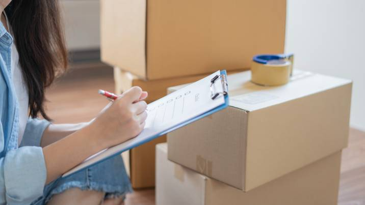 woman writing on moving checklist on clipboard, sitting near moving boxes in living room
