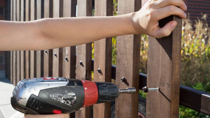 A person's hand operating a power drill to attach wooden boards to a fence in an outdoor setting. - fencing cost