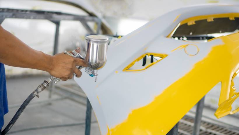 A person spraying yellow paint onto a car bumper using a spray gun. - car respray cost