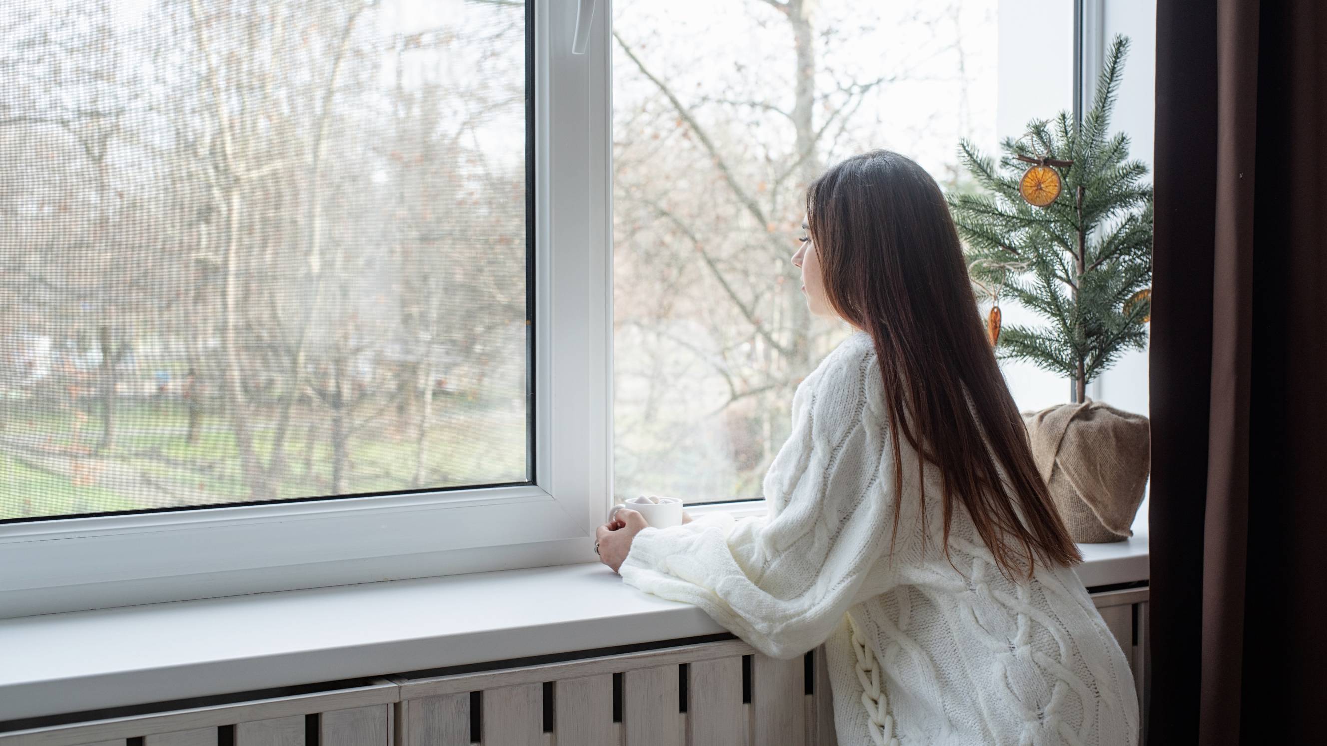 a woman in warm white winter sweater standing next to the window at home at Christmas Eve