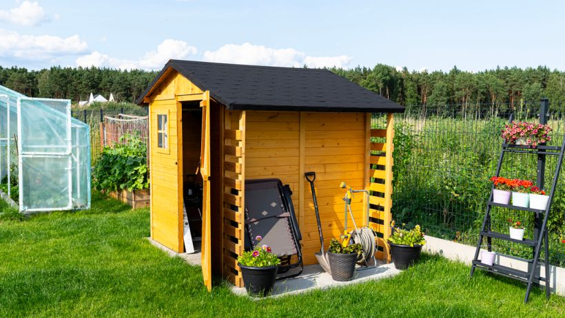 Wooden garden shed with a black roof, positioned in a lush green backyard, storing gardening tools and equipment