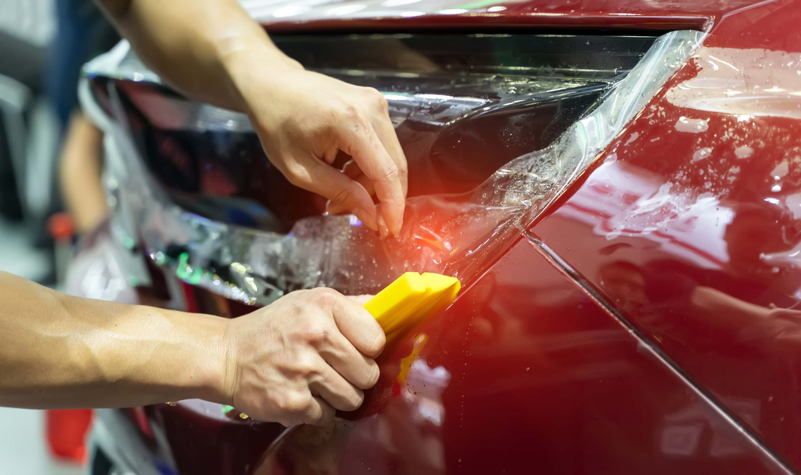 Professional applying car wrap film to a red vehicle's headlight