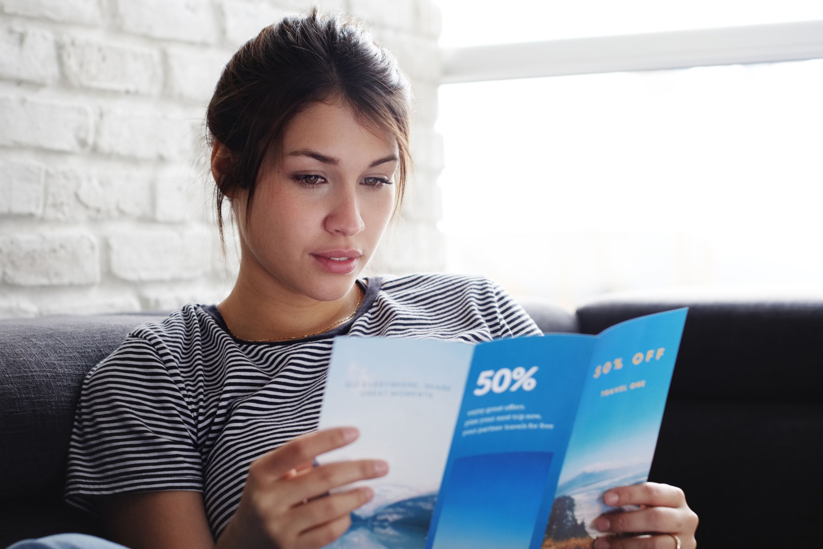 Young hispanic woman laying on couch at home, holding a travel flyer she got from a flyer distributor.