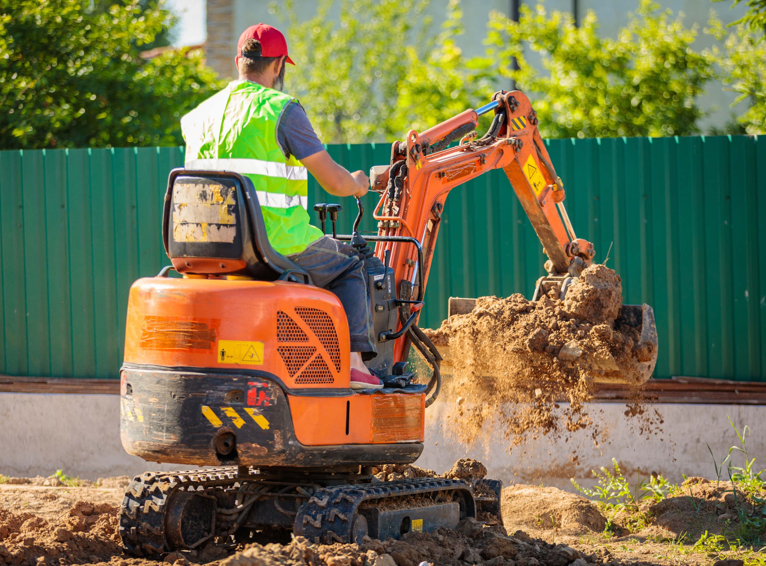 A worker in a safety vest and cap operates a backhoe, scooping soil on a job site.