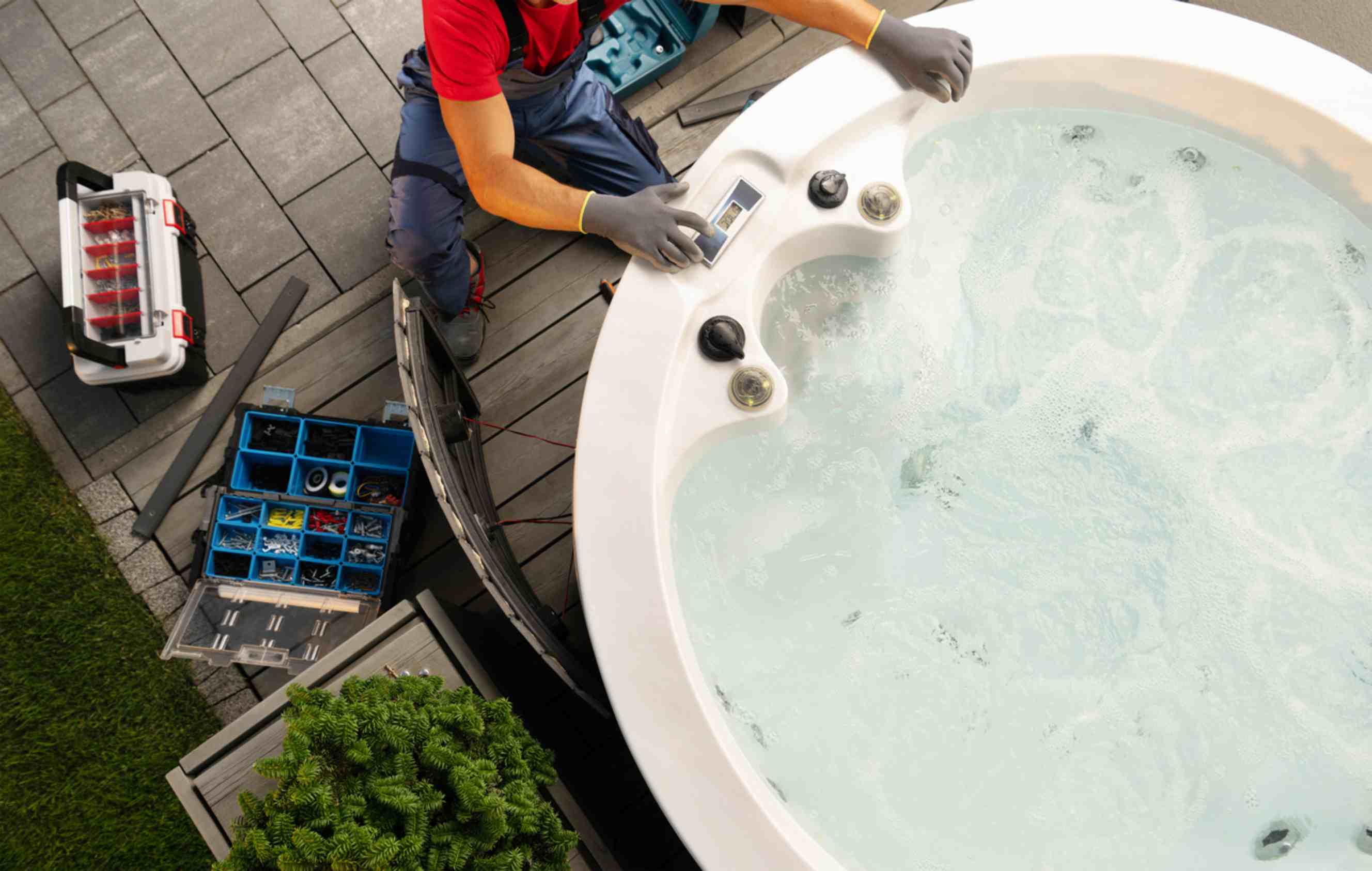 A mover in work gloves and overalls adjusts controls on a large outdoor hot tub while preparing it for moving.