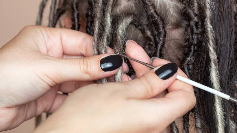 Close-up of hands with black nail polish using a crochet hook for weaving dreadlocks into a client's natural hair. - how much do dreadlocks cost