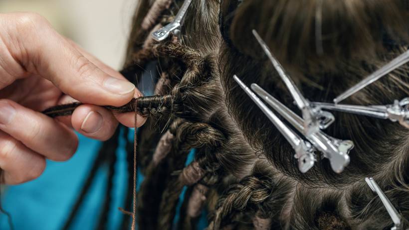 Close-up of a stylist’s hands using brown thread to secure a section while braiding dreadlocks into a client's hair. - dreadlocks cost