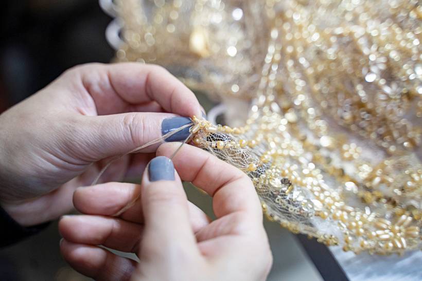 dress alterations cost - Close-up of hands sewing gold beaded lace fabric during dress alterations