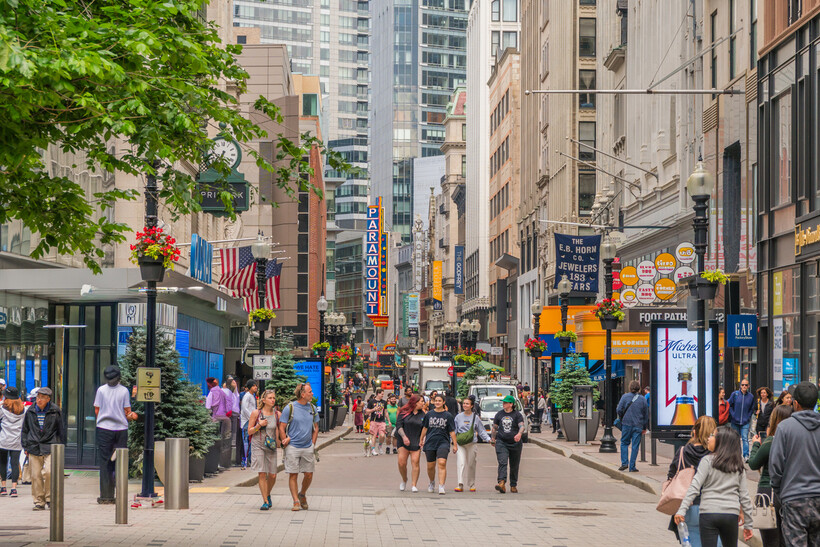 Pedestrians crossing a city street surrounded by retail stores and commercial buildings. - moving to Boston