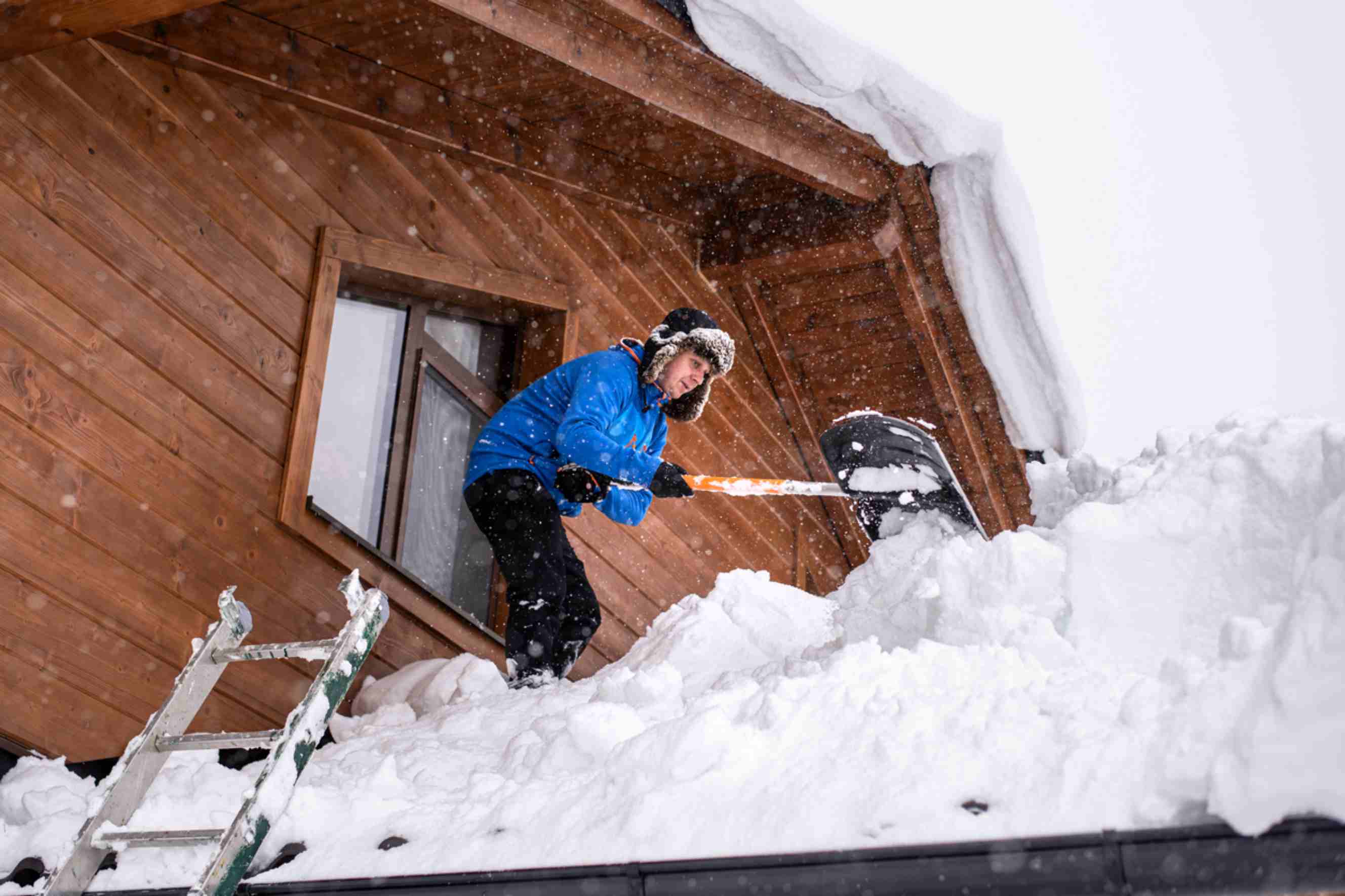 Person performing roof snow removal on a wooden house, standing in deep snow and using a shovel near an open window during active snowfall.