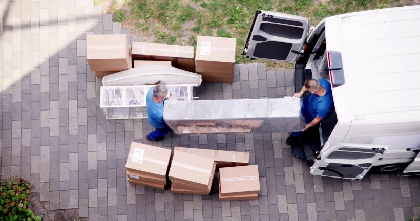 Overhead view of professional movers in blue uniforms loading a plastic-wrapped sofa and cardboard boxes into a white transport van. - moving to las vegas