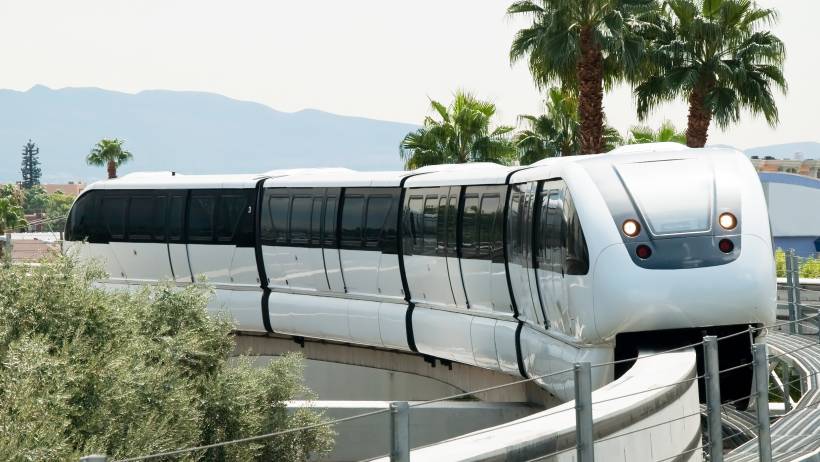 Front view of a modern transit train operating on the Las Vegas Monorail system, surrounded by desert greenery and bright daylight. - moving to las vegas